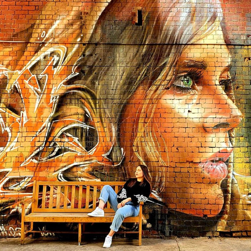 Young woman sitting on a wooden bench in front of a large mural painted on a brick wall